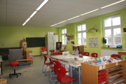 Kind sitzt am Tisch mit roten Stühlen und malt, im Hintergrund ist eine grüne Wand mit Fensterfront, ein großer grauer Schrank, eine Wandtafel und eine Rückzugsecke mit Couch und Sessel, im vorderen Bereich steht ein Materialschrank mit Pinsel, Farben und Schablonen