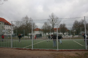 Kinder spielen auf dem Bolzplatz des Hortes Fußball, Bolzplatz besteht aus Eisenstangenzaun und Auffangnetzen an den Seiten,Schulgebäude,Bäume und Sitzmöglichkeiten im Hintergrund 