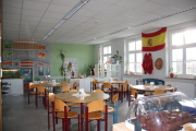 Raum mit 7 Tischen und zugehörigen Stühlen,große Fensterfront, grüne bemalte Wand mit Markise, Plakat, Fisch-Aquarium, Schrankreihe und Bank im Hintergrund, im Vordergrund steht eine gefüllte Kuchenbox auf dem Tresen, Gesundheits-und Körper Spielmaterialien im Raum verteilt