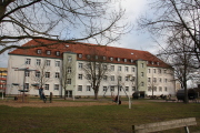 Hort Grabow Gebäude Haus C von Außen fotografiert, kleiner Blick auf den Schulhof, Kinder klettern auf dem Klettergerüst, Kinder schaukeln mit der Vogelnestschaukel,Kinder sitzen auf der Bank und laufen über den Hof, 2 Sitzmöglichkeiten mit Dach, 8 Bäume verteilt aus dieser Ansicht, Gebäude ist weiß mit grün Bildunterschrift: Hort Grabow Gebäude 