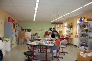 Großer Raum mit viel Bastelmaterial, Kinder sitzen verteilt an drei Tischen und basteln, an der linken Wand steht ein Schreibtisch und eine große Schrankwand, Pappkartons und anderes Material, an der rechten Wand ist eine Fensterfront und ein großer Schrank gefüllt mit Bastelmaterial