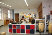  Musikraum mit Sicht auf die Instrumentenecke, Gitarren, Bongos, Cajons; Boomwhakers, Kongas sind zu sehen, im Vordergrund steht ein Regal auf dem Boden bestückt mit Stoffeinsätzen, Schallplatten, Musikanlage, Plattenspieler und Mischpult, im Hintergrund sitzt ein Kind am Tisch und malt nach Musik 