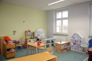 Bereich des Spielzimmers in der Teppichecke mit Kaufmannsladen, Küche und Zubehör, in der Mitte ein Tisch mit 2 Stühlen, an der Fensterwand steht ein weißes Puppenhaus mit Zubehör, an der linken Seite stehen noch 2 kleine Schränke mit Puppenspielzeug 