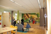 Kinder spielen auf der Bühne, einige sind verkleidet, einige schauen nur dem Geschehen zu, im Vordergrund Tisch mit 2 Stühlen, im Hintergrund großer Schrank, Vorhänge und Theaterbühnenhintergrundbild