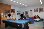 2 Kinder spielen am Billardtisch mit Kugeln, im Hintergrund sitzen Kinder auf der Couch und unterhalten sich, im Hintergrund ist auch ein Tresen mit Hochstühlen, ein Regal mit Spielen und eine Schrankwand zu sehen, an der grau-blauen Wand hängt eine Dartscheibe mit Styropor, daneben hängen Plakate  